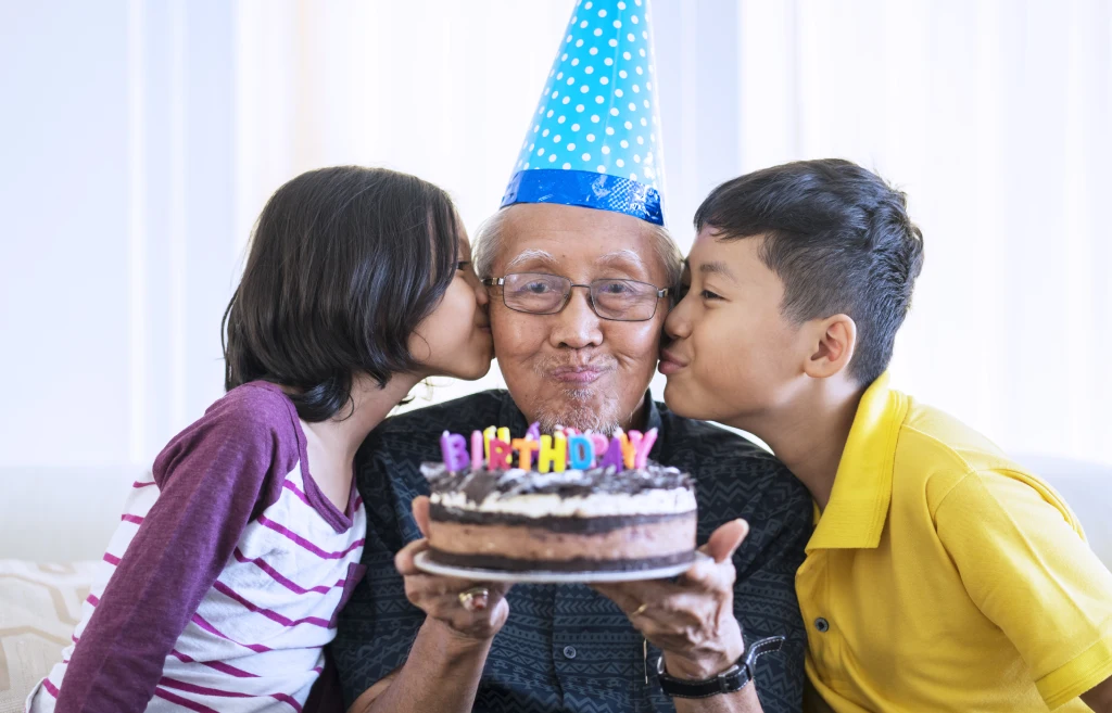 Elderly man holding a birthday cake, kissed by two children